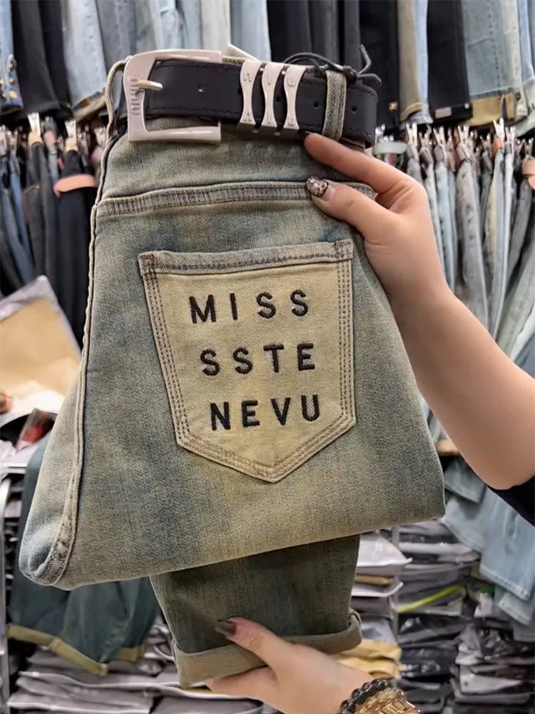 Denim pants with a pocket displaying 'MISS SSTE NEVU' held in front of a clothing rack.